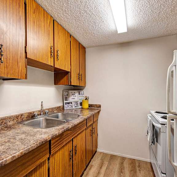 narrow kitchen here at lakewood heights apartment homes with brown wood cabinets, marble countertops, a double sink, and white appliances. bright, clean, and simple atmosphere.