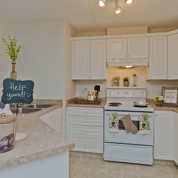 cozy kitchen here at somerset pointe apartment homes with white cabinets, a stove, and decor items like plants and framed art. a "help yourself" sign sits on a counter with a candy jar.