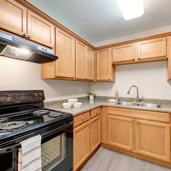 a cozy kitchen here at wyndham crossing apartment homes with light wood cabinets, granite countertops, and a stainless steel sink. a black stove and fridge add contrast. bright and tidy ambiance.