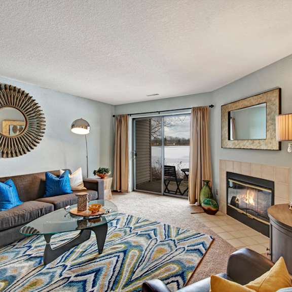 a bright living room here at river’s cove with a gray sofa and blue accent pillows, patterned blue rug, and cozy fireplace. large sliding doors bring in natural light, creating a warm, welcoming atmosphere.