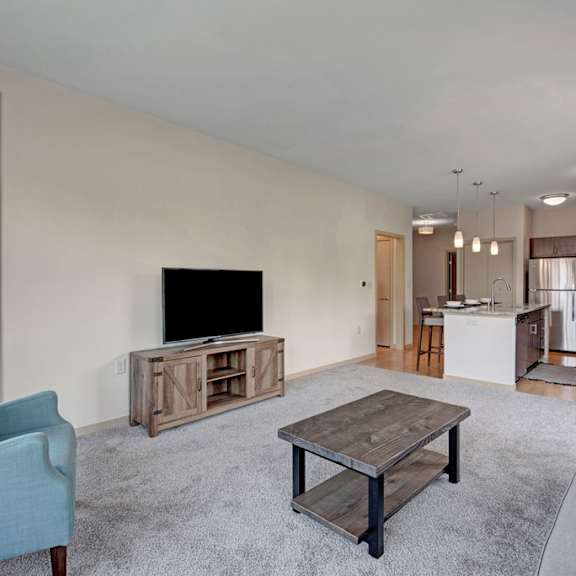 a bright living room here at vue at pinnacle park with soft carpet, a gray sofa, teal accent chair, wood coffee table, tv console, and an open kitchen with pendant lights and warm wood cabinets.