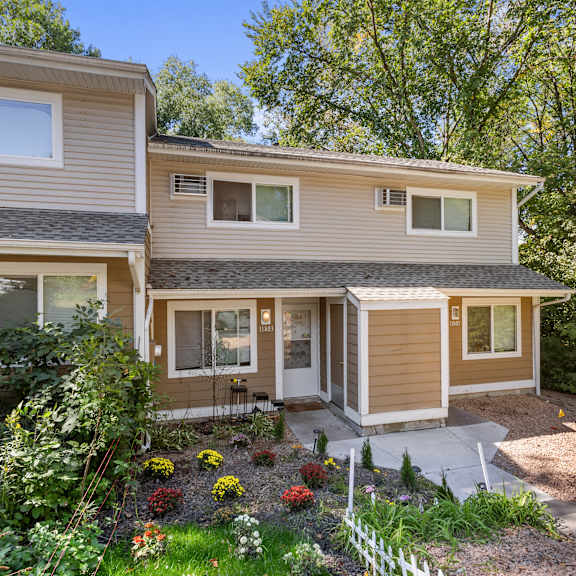 a house with a brown and beige exterior is surrounded by greenery and has a small garden in front.