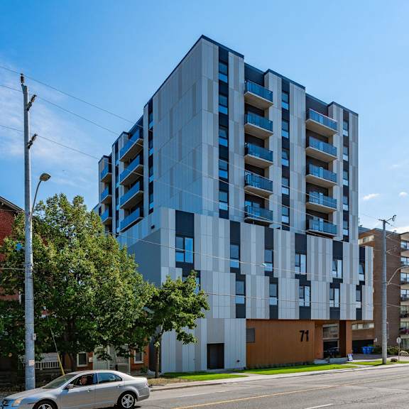 a modern multi-story building with blue and white balconies is situated on a street corner with a car and a cyclist in the foreground.