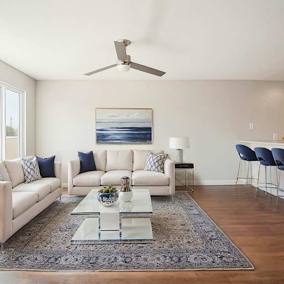 a modern living room with a white sofa set, a glass coffee table, and a ceiling fan.