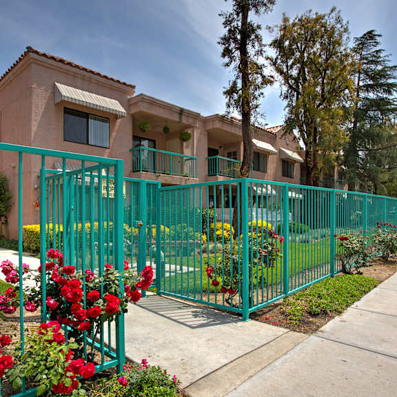 a green fence surrounds a building with red flowers in front.