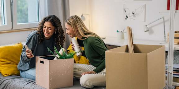 Two women are sitting on a bed with boxes and a plant, possibly moving in or out of the home.