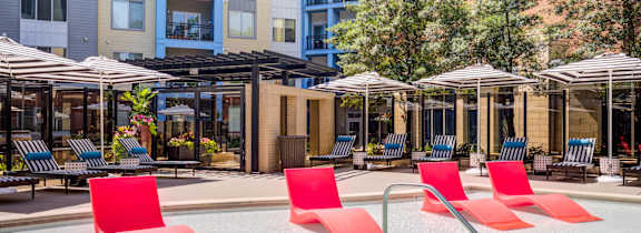 A pool with red chairs and umbrellas in front of apartment buildings.