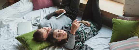 A couple is lying on a bed with pillows and a laptop.