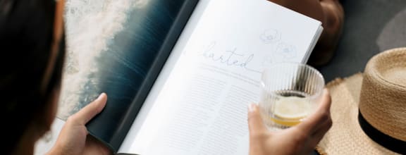 a woman reading a book while holding a glass and a straw hat
