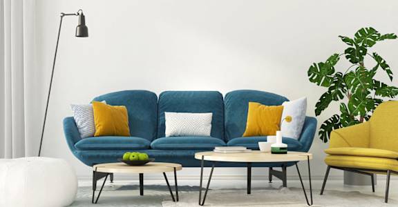A blue sofa with yellow pillows sits in a room with a white floor lamp and a white table with a green plant on it at Ballantyne Commons of Stockbridge Apartments, Georgia