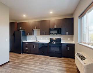 A kitchen with dark wood cabinets next to a living area with hardwood floors and a large window