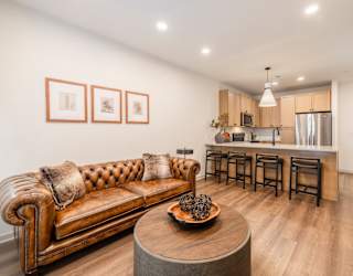 A brown leather couch with pillows sits in a living room with a kitchen in the background.