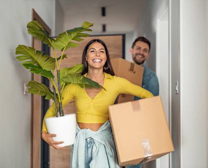 A woman is holding a potted plant and a cardboard box while a man is carrying another cardboard box.