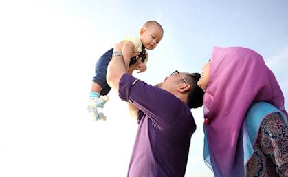 A man in a purple shirt is lifting a baby up in the air.