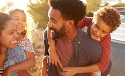 A man is carrying a child on his back while another child looks on.