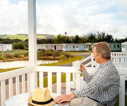 A man in a plaid shirt and hat sits on a porch with a cup in hand.