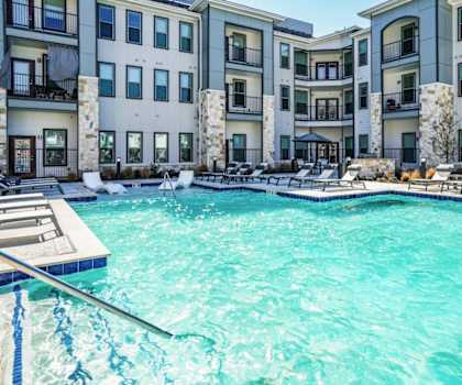 A swimming pool in front of a building with a deck and lounge chairs.