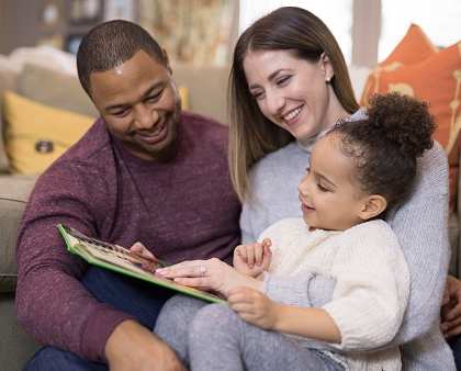 A family of three is sitting on a couch, reading a book together.