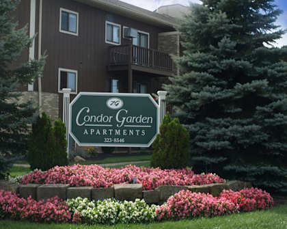 property sign with large evergreen trees on either side and a brown apartment building in the background