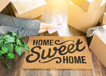 A person is unpacking boxes on a doormat that says "Home Sweet Home".