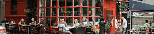 A red building with a black awning and people sitting at tables outside.