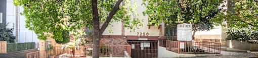 Tree lined property entrance with brick and metal gate at Joro Properties Apartments, Hollywood, CA 90046