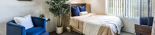 A bedroom with a bed, a blue chair, and a small tree.