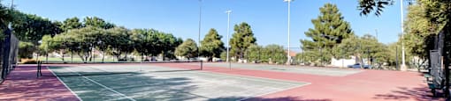 A tennis court with a red surface and white lines, surrounded by trees and a clear blue sky.