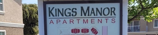 A Kings Manor Apartments sign is in front of a tree and a building.