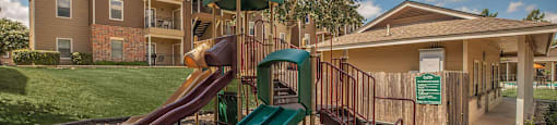 A playground with a green slide and a red slide in front of a building at The Villages at Lost Creek Apartments, San Antonio, TX