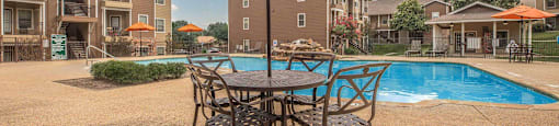 A patio with a table and chairs under an umbrella with a pool and apartment buildings in the background at The Villages at Lost Creek Apartments, Texas, 78247