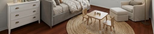 A cozy bedroom with a grey bed, a white dresser, a wooden coffee table, and a beige armchair.