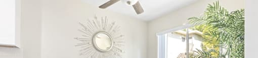 Encino, CA Apartments - Brookside Terrace - Bright Dining Nook with a Table, Four Chairs, a Ceiling Fan, White Walls, a Houseplant, a Mounted Mirror with a Sun Design, and a Large Window