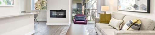 Apartments in Encino, CA for Rent - Brookside Terrace - Spacious Living Room Area with White Sofa, Rug, Wooden Coffee Table, Wood-Style Flooring, Mounted Art, Balcony Access, Large Windows, Purple Armchair, and Electric Fireplace