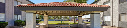 A pavilion with a red tile roof is situated in a grassy area in front of a building.