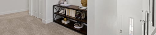 A hallway with a carpeted floor, a mirror, a framed picture, a console table with a vase and books, and a staircase.