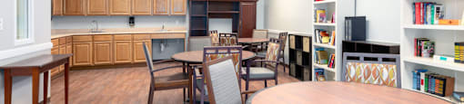 A room with a wooden table and chairs, a bookshelf, and a kitchen area in the background.