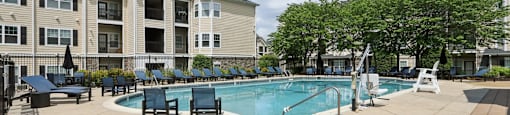 A large swimming pool surrounded by lounge chairs and trees in front of apartment buildings.