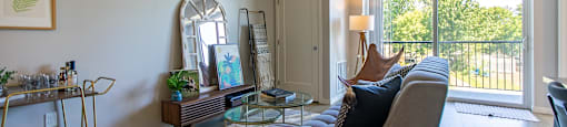 A living room with a grey couch, a glass table, and a large window.
