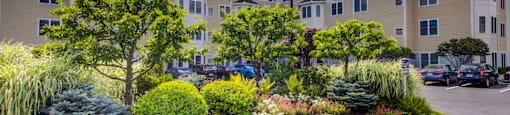 View of community with lush and colorful landscaping with apartment building parking lot of cars