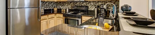 U-shaped kitchen with black countertops, stainless steel refrigerator, microwave and stove. Ample cabinet space with blonde cabinets above and below counter with multi-colored tile backsplash