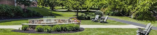 Fire pit with built-in bench seating surrounded by ample green space and trees with part of building in the background.