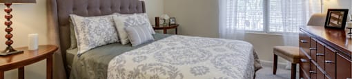 Furnished bedroom with plush carpeting and oversized window with mini-blinds. Room includes queen-sized bed, two nightstands, chair, lamp and horizontal dresser. at Summit Place apartments in Methuen,