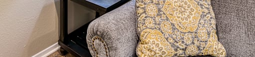 A grey couch with a yellow and white pillow sits next to a table with a lamp and candle.