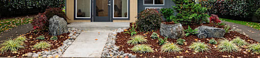 A house with a grey exterior and a brown door is surrounded by a garden with red and green plants.