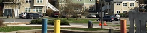 A park with a bench, a trash can, and a large letter "D" in the foreground with houses in the background.