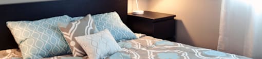 A bed with a grey and white patterned comforter and two blue patterned pillows sits in a room with a framed picture on the wall and a lamp on a side table.