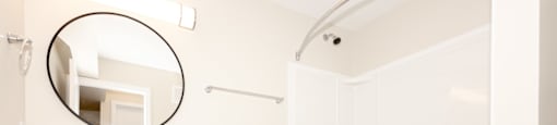 A white toilet is in a bathroom with a white tub and a round mirror.