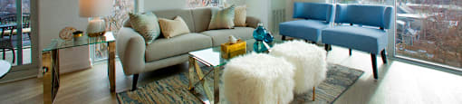 Living room surrounded by large windows overlooking the nearby scenery. There are two fluffy white stools and two blue chairs in the center of the room