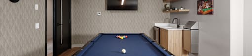 A pool table is in the middle of a room with a television and a bar in the background at The Ambrosi in Kelowna, BC.